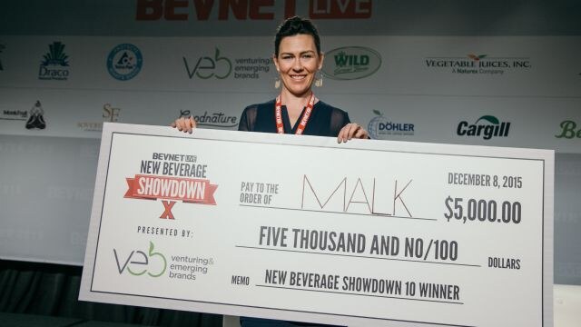 A woman with dark hair and a lanyard holds a large check made out to MALK for $5,000, marked New Beverage Showdown 10 winner, in front of a sponsor-logo backdrop.