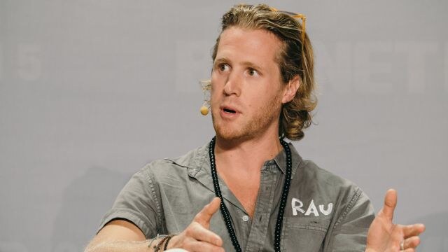 A man with shoulder-length blond hair and a headset microphone gestures with both hands while speaking. He wears a gray button shirt with RAU printed on it and a black beaded necklace.