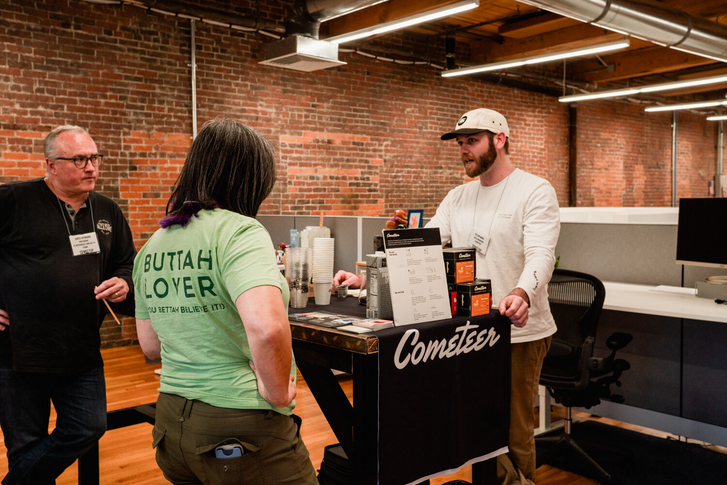 Several people stand in the brick-walled office around a black table branded Cometeer, with product boxes on top.