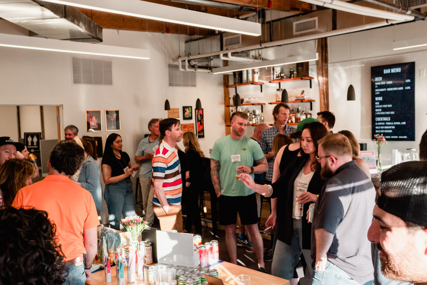 Wide shot of guests talking in a bright brick-walled office with a bar, shelves of bottles and a bar menu screen.