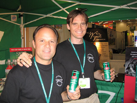 Two men in black Sjeez shirts and green lanyards smile and each holds a green can under a green canopy, with a Wizards Cauldron Inc. sign behind them.