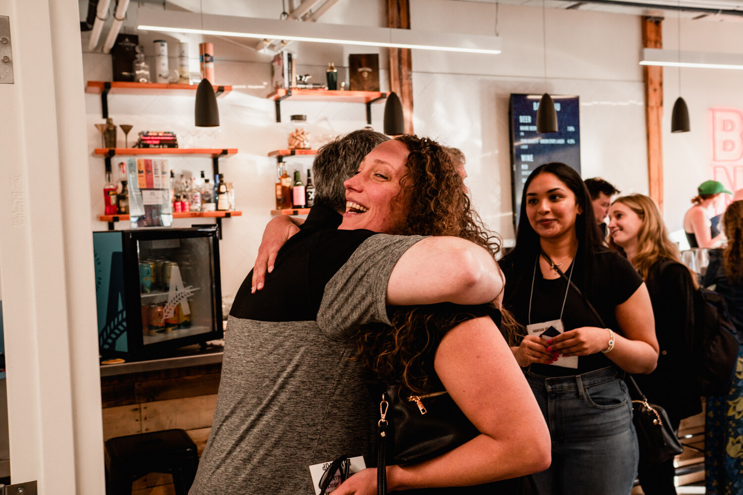 A man in a gray polo and a woman in a black top hug and laugh in front of a bar with shelves of bottles.