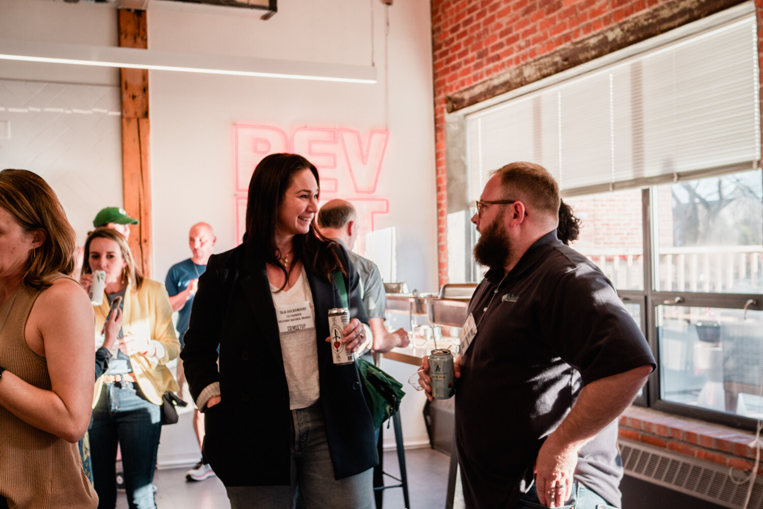 A smiling woman in a dark blazer and a bearded man in a polo hold drinks and talk near a window and pink BEVNET neon sign.