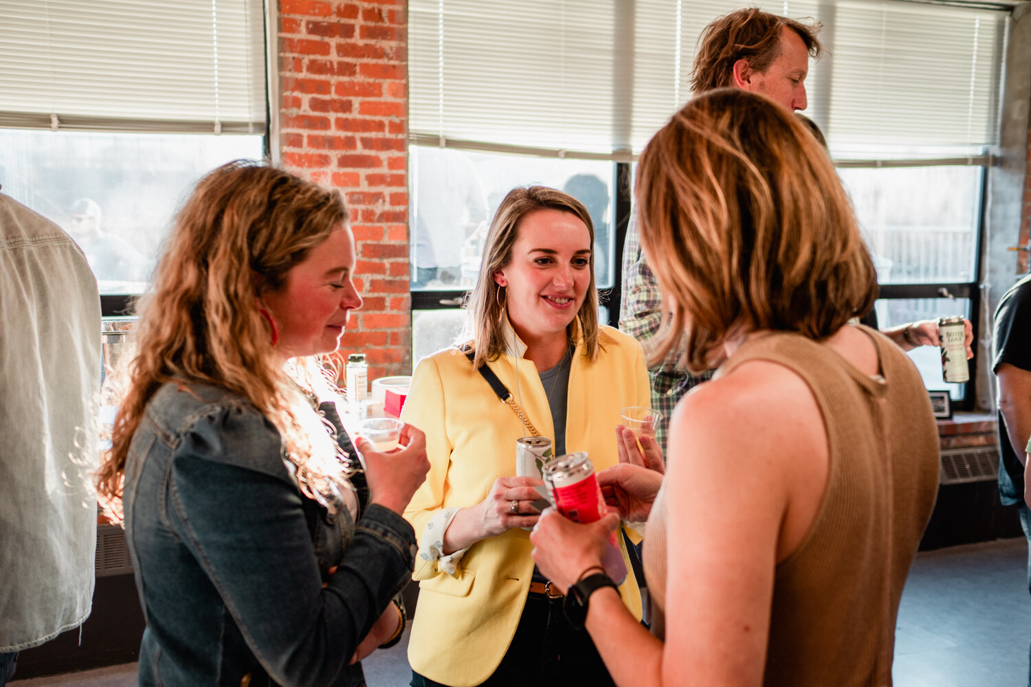 Three women hold cans and small cups together near a bright window, the one in the middle wearing a yellow blazer and smiling.