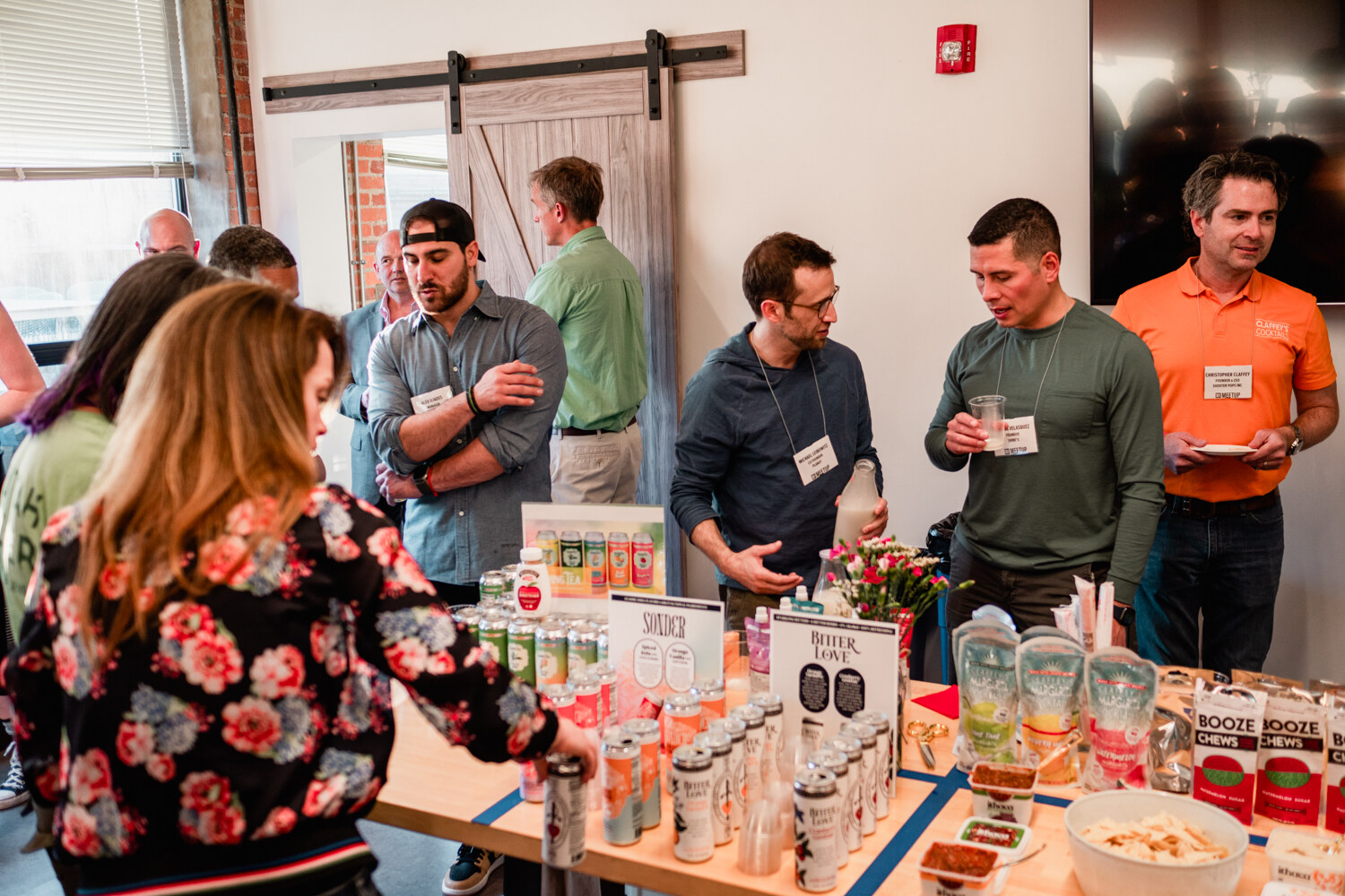 Several men stand behind a table crowded with canned drinks, Booze Chews and flowers near a sliding barn door.