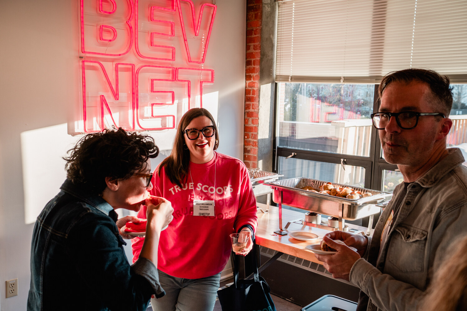 A woman in a red True Scoop sweatshirt talks with a person in a denim jacket and a man in glasses, with a BEVNET neon sign behind them.