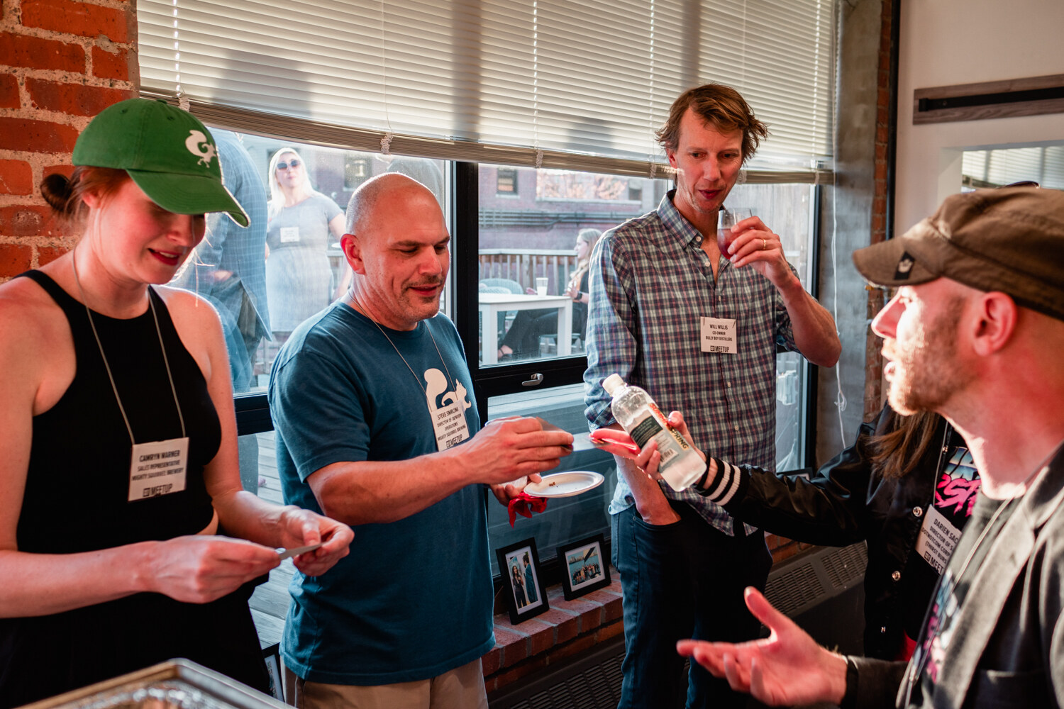 Four guests wearing name badges exchange cards in front of a window with a plate of food in hand.