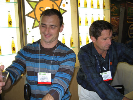Two men wearing name badges sit in front of shelves of yellow bottles and a sun logo with the word Sol. One man in a striped sweater looks down and smiles; the other wears a dark plaid shirt.