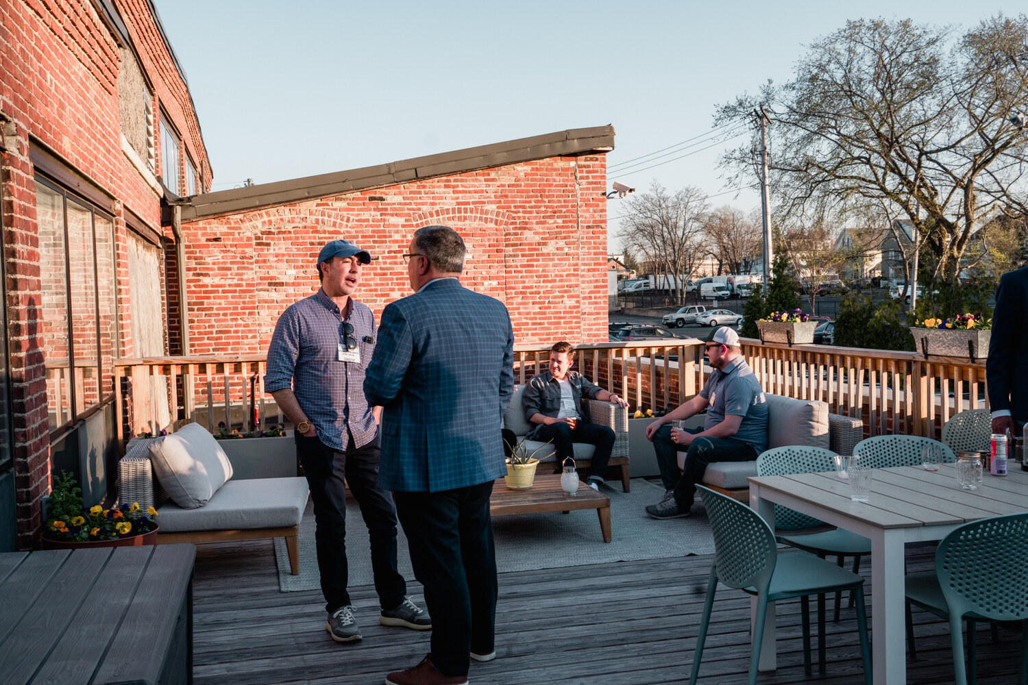Two men in button-up shirts talk on a wooden deck with outdoor furniture and a brick wall behind them.