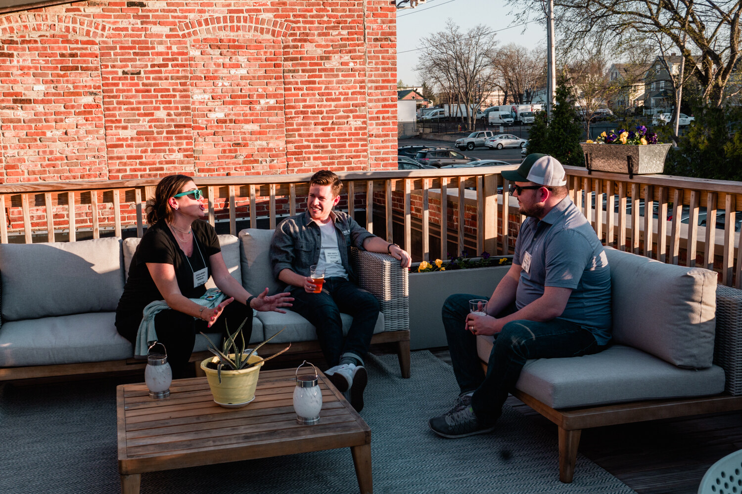 Three guests with drinks sit on gray outdoor sofas on a deck beside a brick wall.