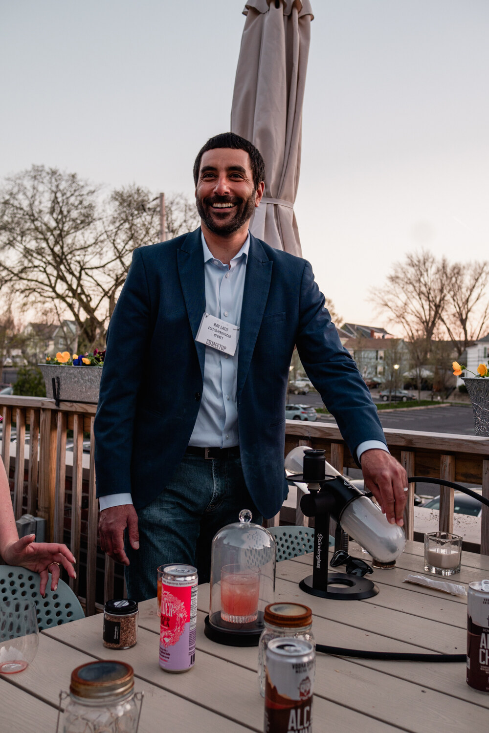 A smiling man in a navy blazer and name badge works with a cocktail smoker on a table with cans and glasses as dusk falls.