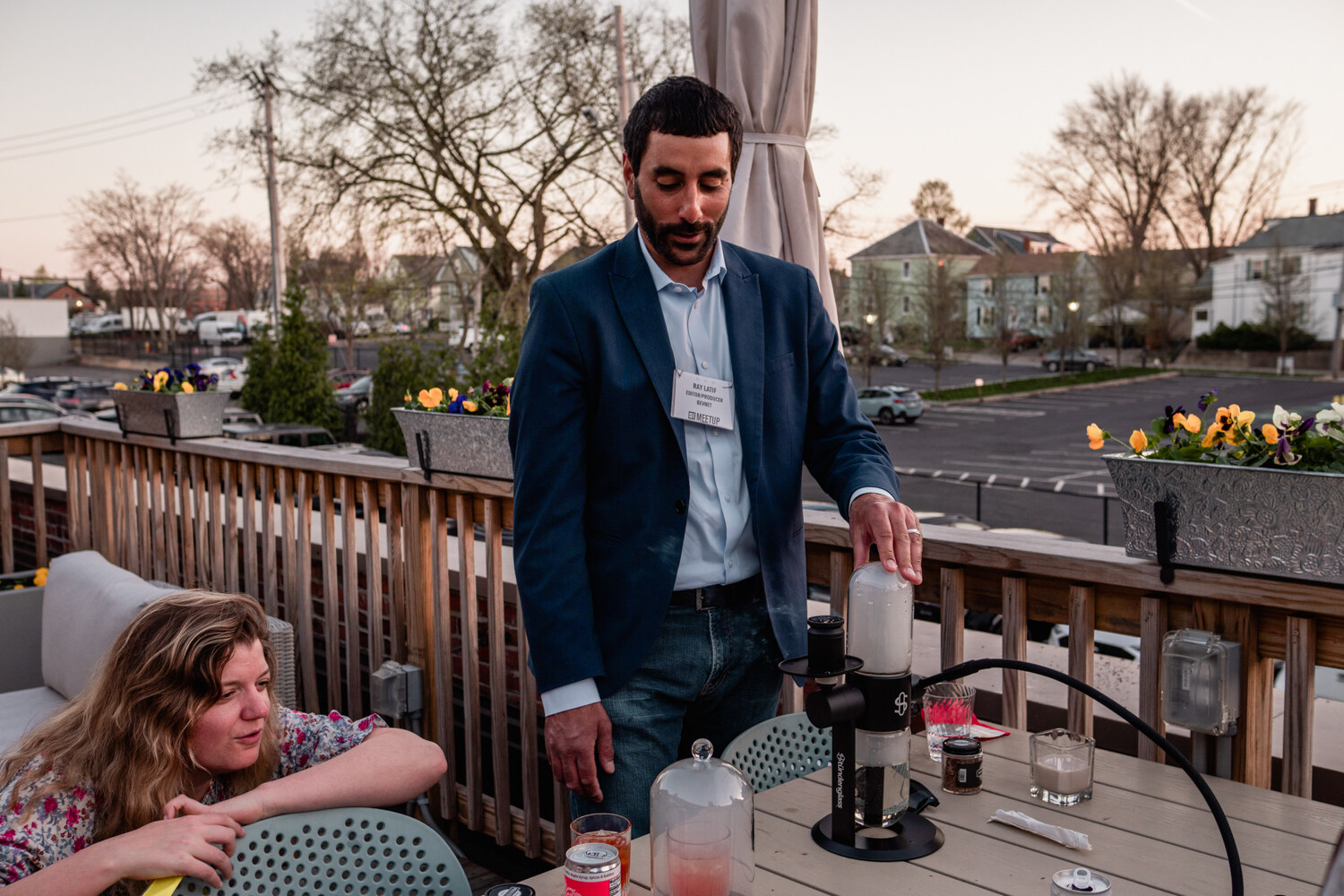 A man in a navy blazer prepares a drink with a smoking device on a deck table, with a seated guest in the corner.