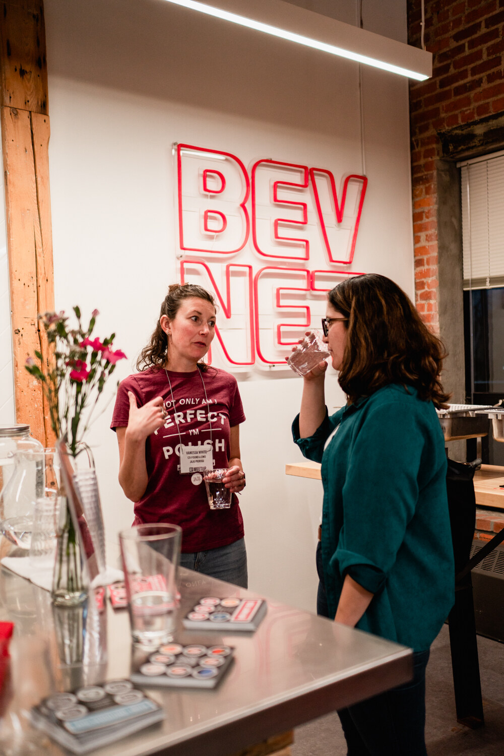 Two women talk beside a bar counter with flowers in front of a pink BEVNET neon sign.