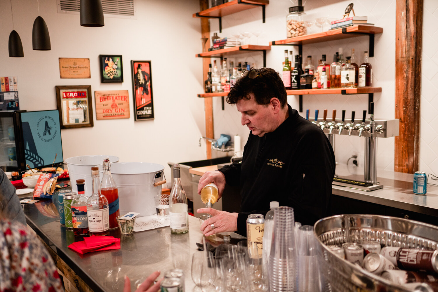 A man in black pours a drink behind a steel bar with shelves of liquor and a row of taps behind him.