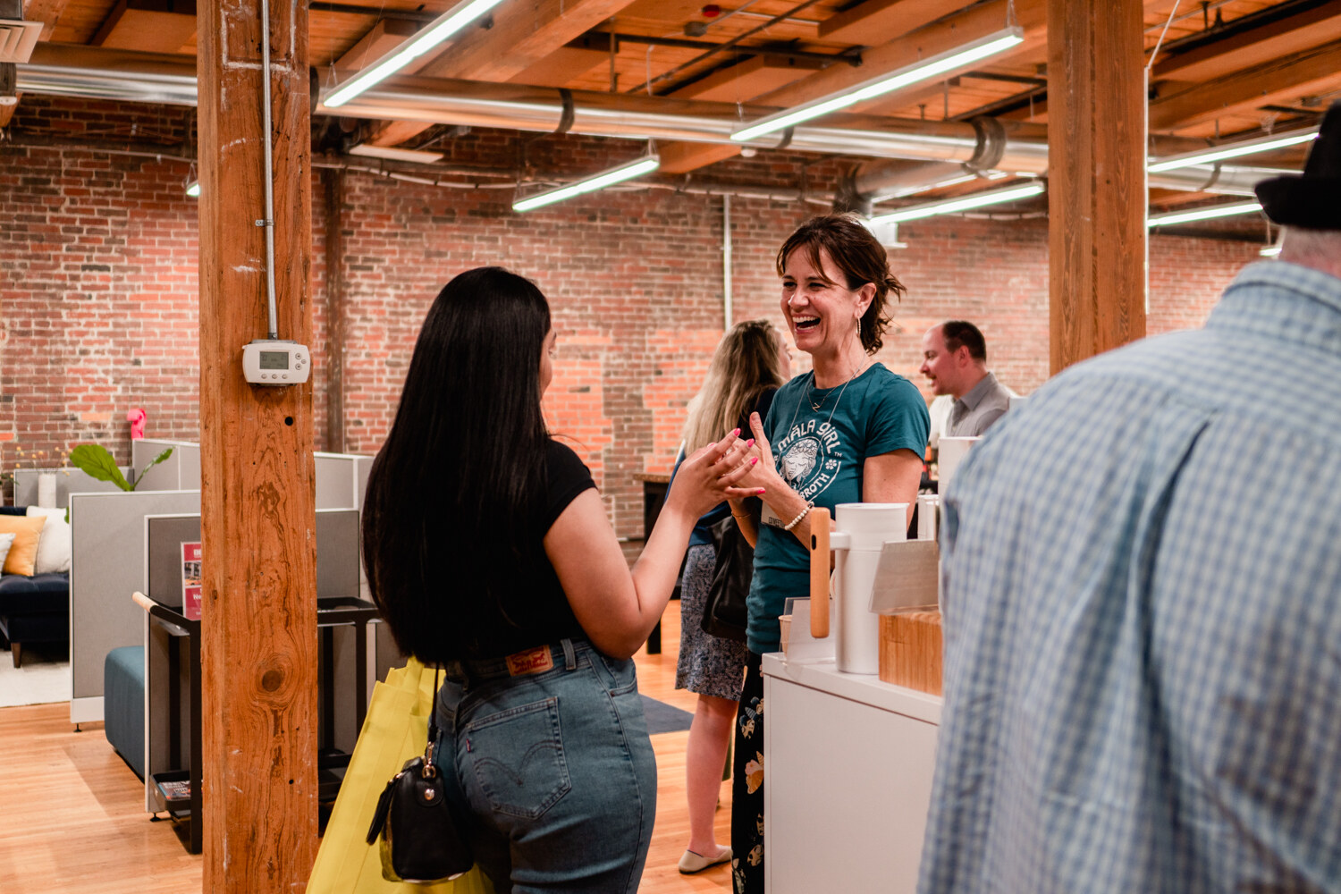 A smiling woman in a teal shirt talks with a woman in black while guests stand in the open office.