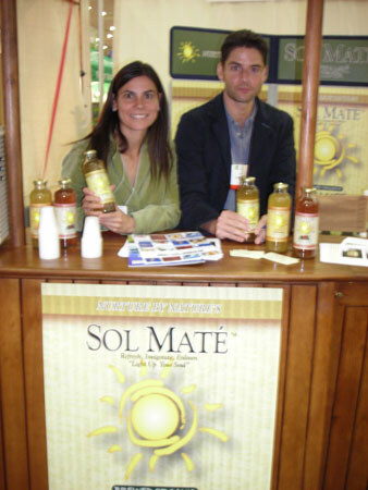 A woman and a man sit behind a wooden counter holding bottles of Sol Maté, with brochures on the counter. A Sol Maté sun logo banner hangs on the front of the counter.
