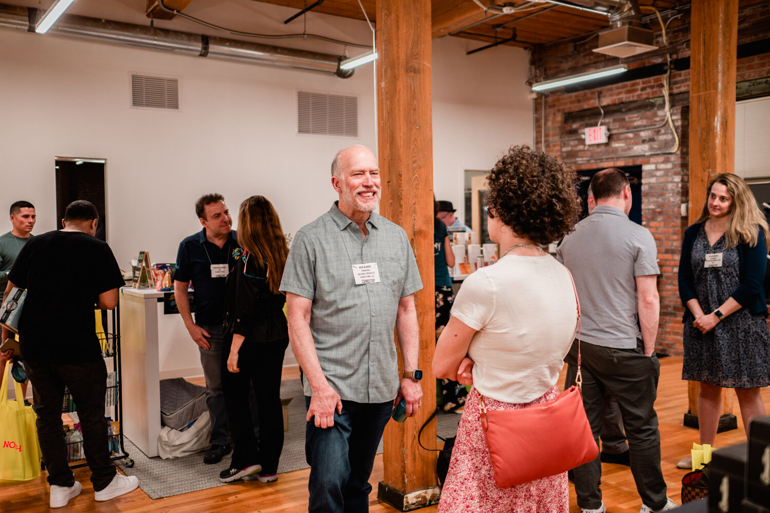 A smiling man in a gray shirt talks with a woman in a cream top and others in the brick-walled office.