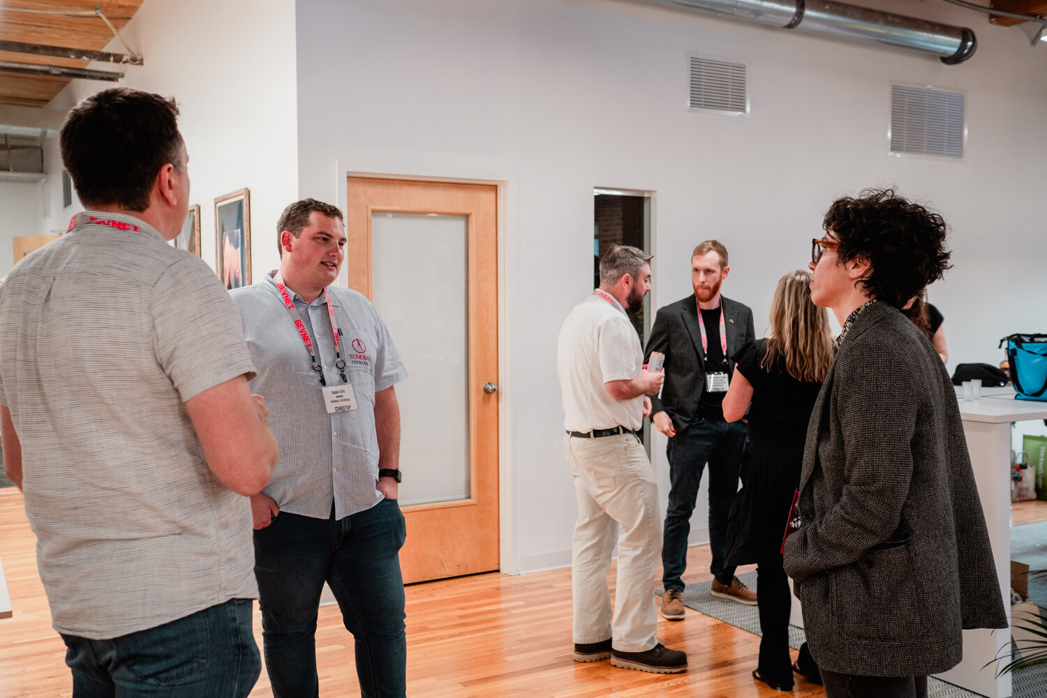 Several guests wearing BevNET lanyards stand and talk in a bright office with wood doors and bottles on a table.