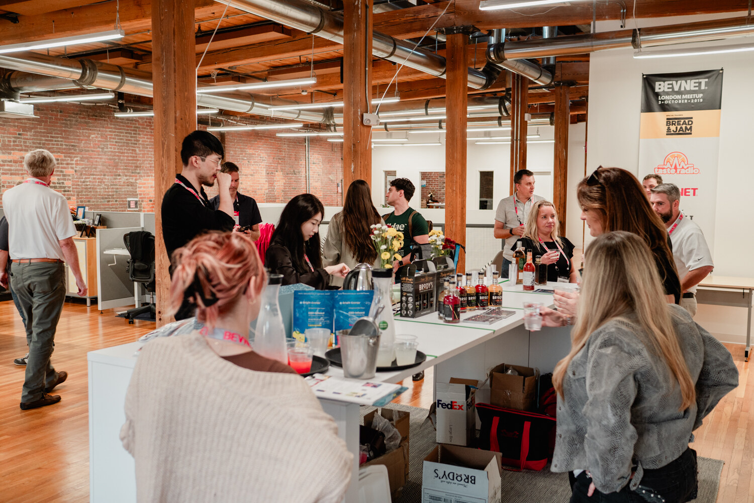 Guests gather around white tables with sampling items in a brick loft with a BevNET banner behind them.