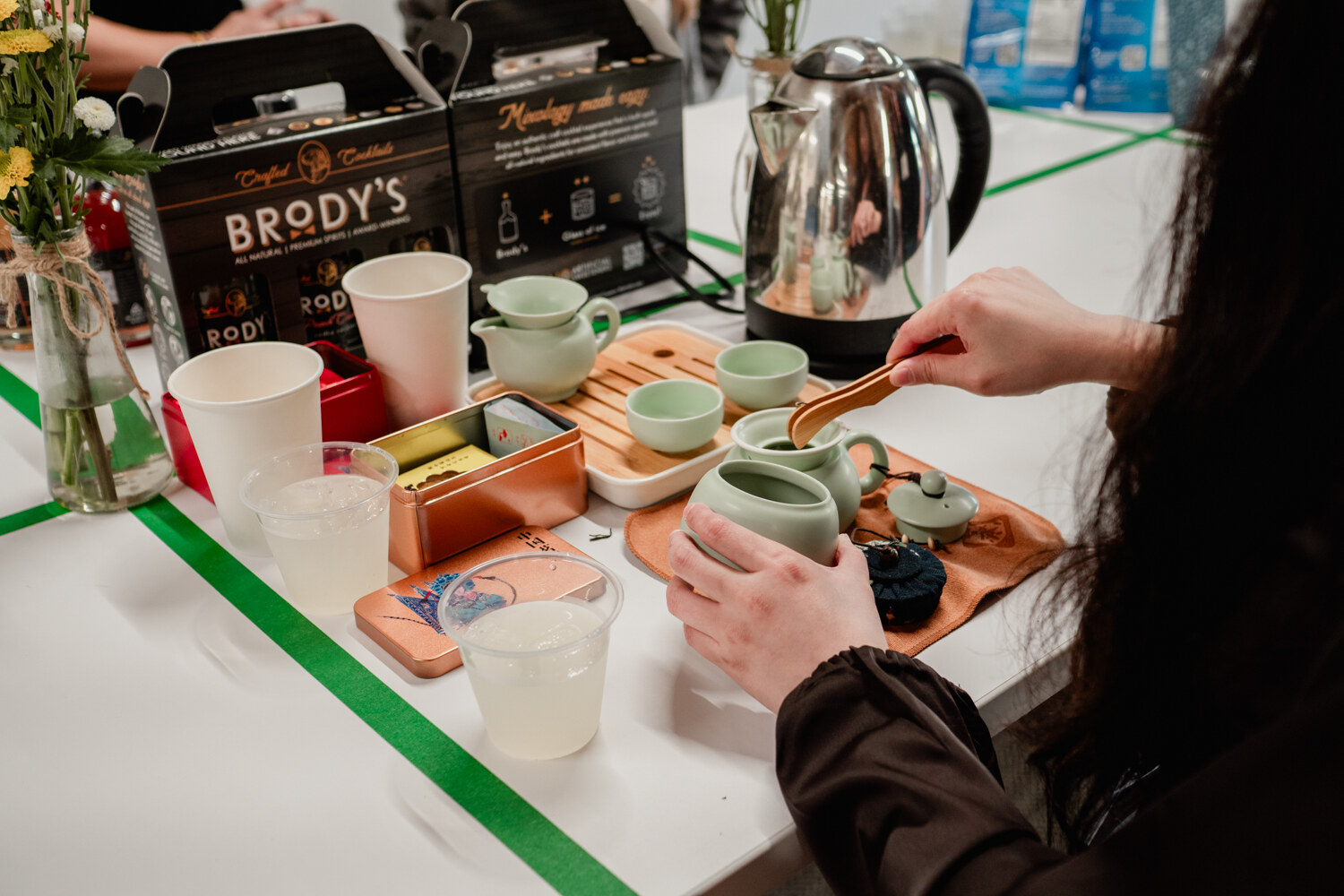 A hand pours tea over a tray of small cups near boxes labeled Brody's and an electric kettle.