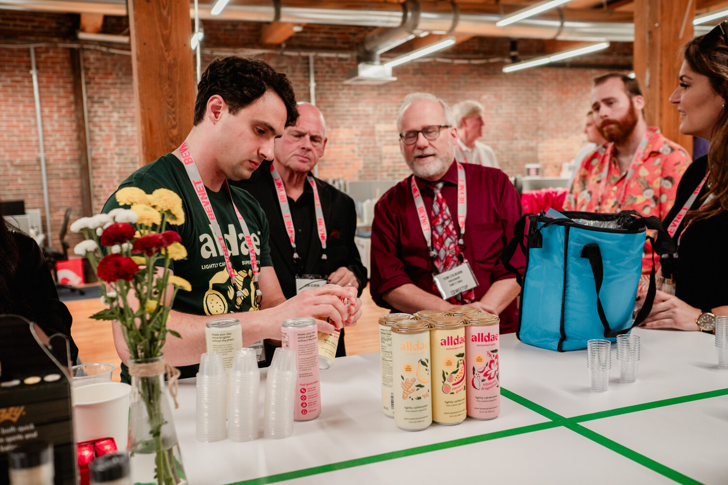 A man in a green Alldae shirt talks with two men in lanyards over a table with canned drinks and a blue cooler bag.