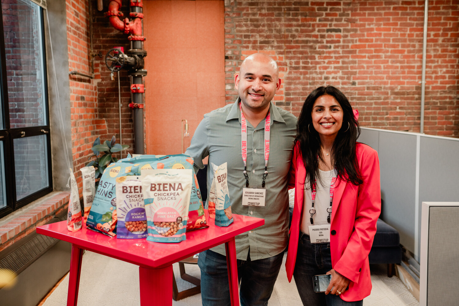 A man and a woman in a pink blazer pose together beside a red table stacked with Biena chickpea snack bags.