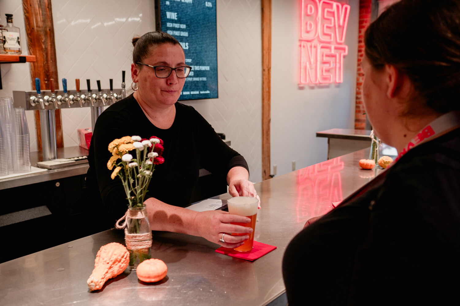 A woman at a steel bar pours a drink into a glass with the pink BEVNET neon sign glowing behind her.
