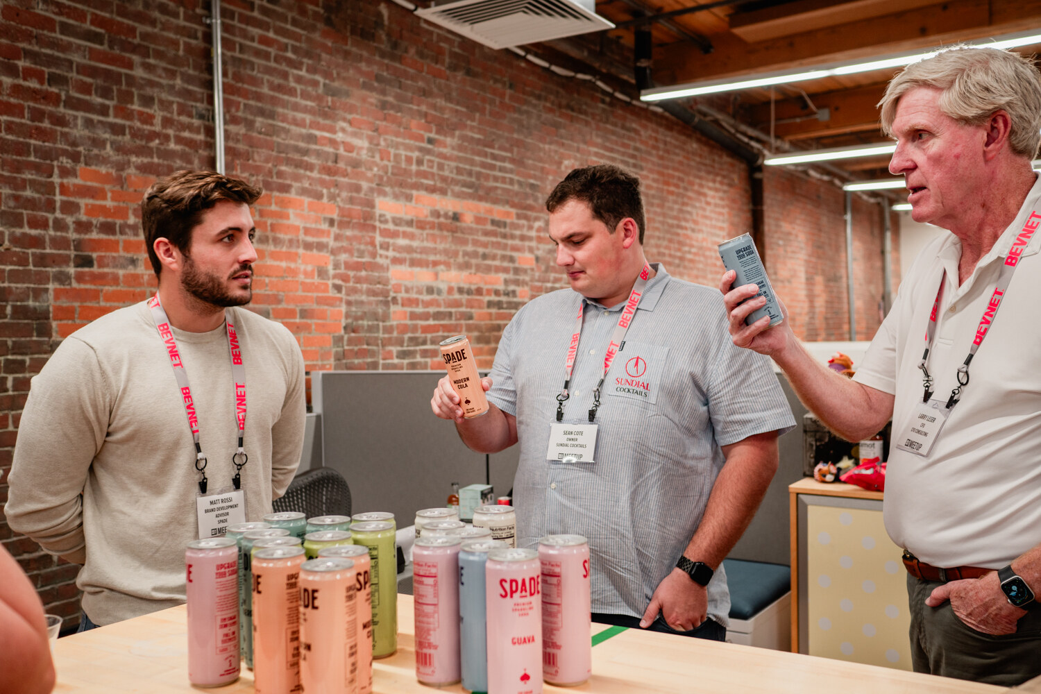 Three men with lanyards talk and hold cans behind a table of Spade sparkling soda cans in several colors.