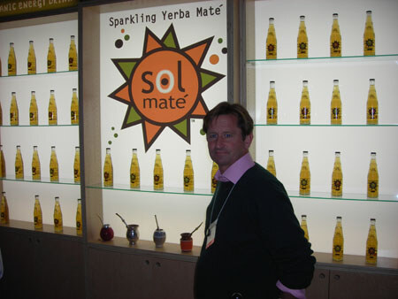 A man in a dark sweater and badge stands beside a lit shelf wall of Sol Maté sparkling yerba maté bottles, with a large orange Sol Maté sun logo and several gourd cups on a shelf.