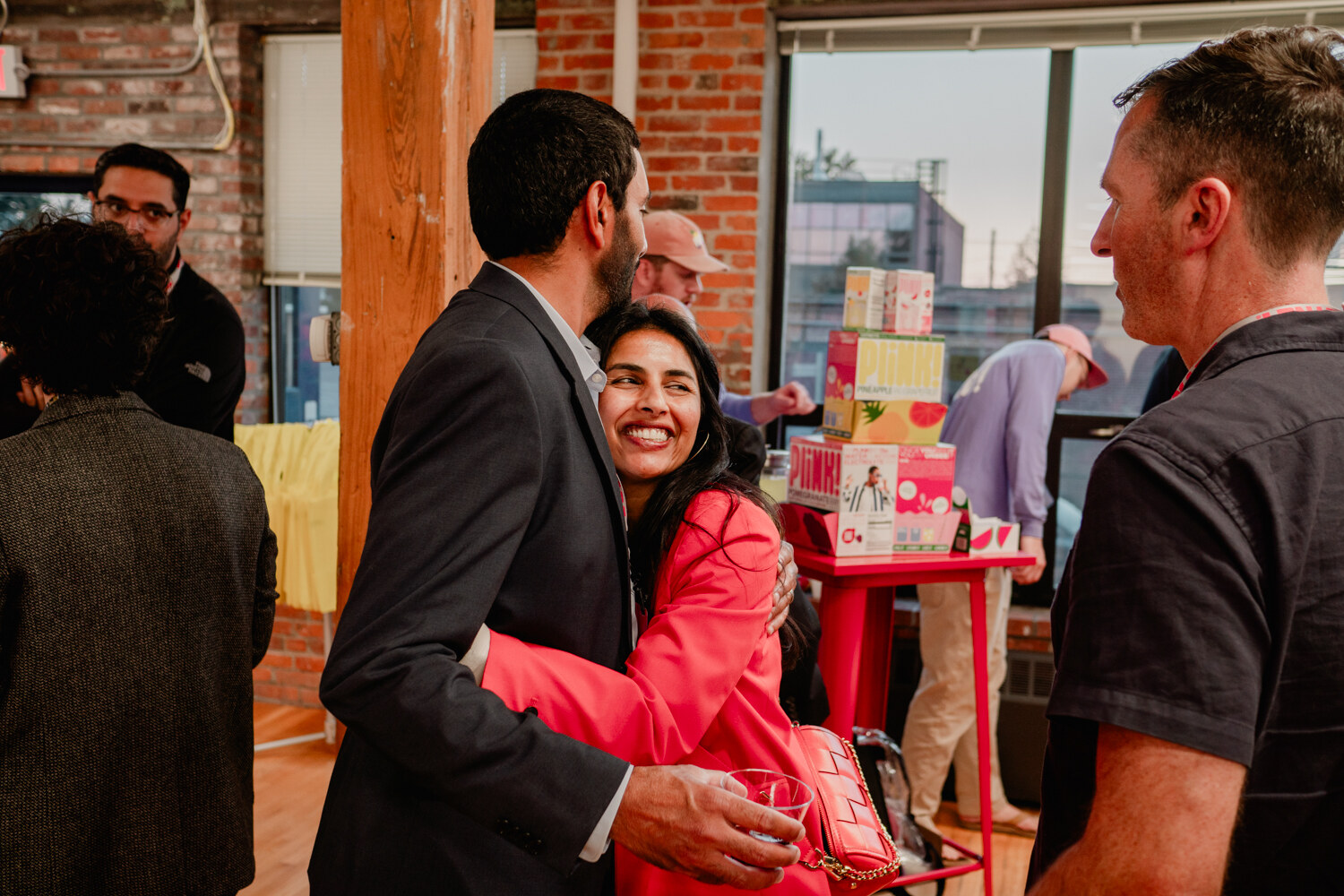 A woman in a bright pink blazer and a man in a dark suit hug while another guest looks on.