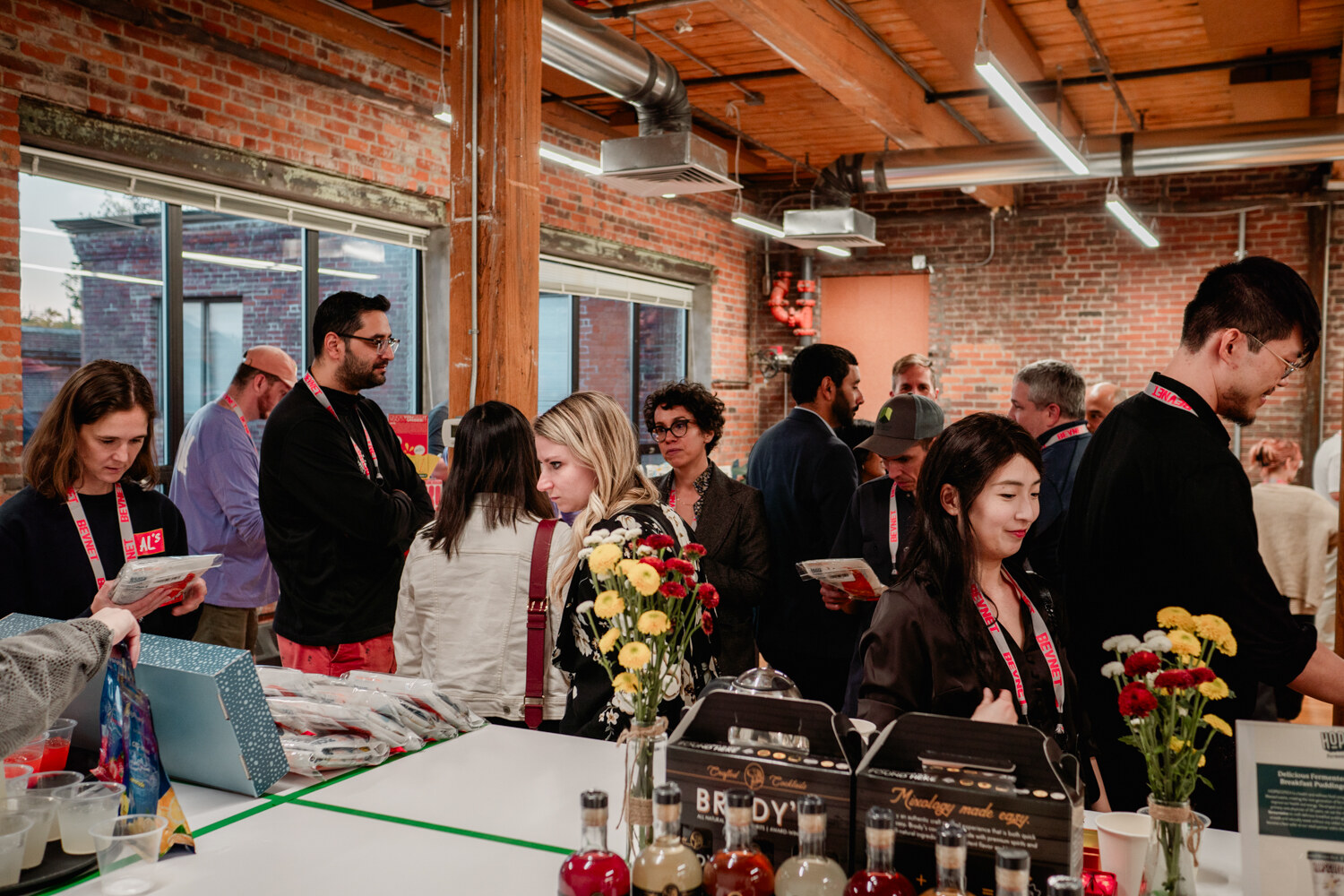 Guests with lanyards eat and talk around a table of Brody's boxes and bottles, with flowers in the foreground.