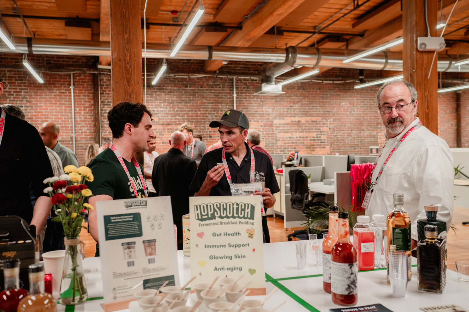 Two men talk with a man in a white jacket behind a table with Hopscotch fermented breakfast pudding signs and sample cups.