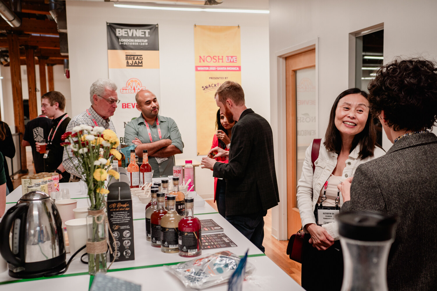 A smiling woman with a lanyard talks with another guest at a table with bottles of Brody's mixers, flowers and an electric kettle.