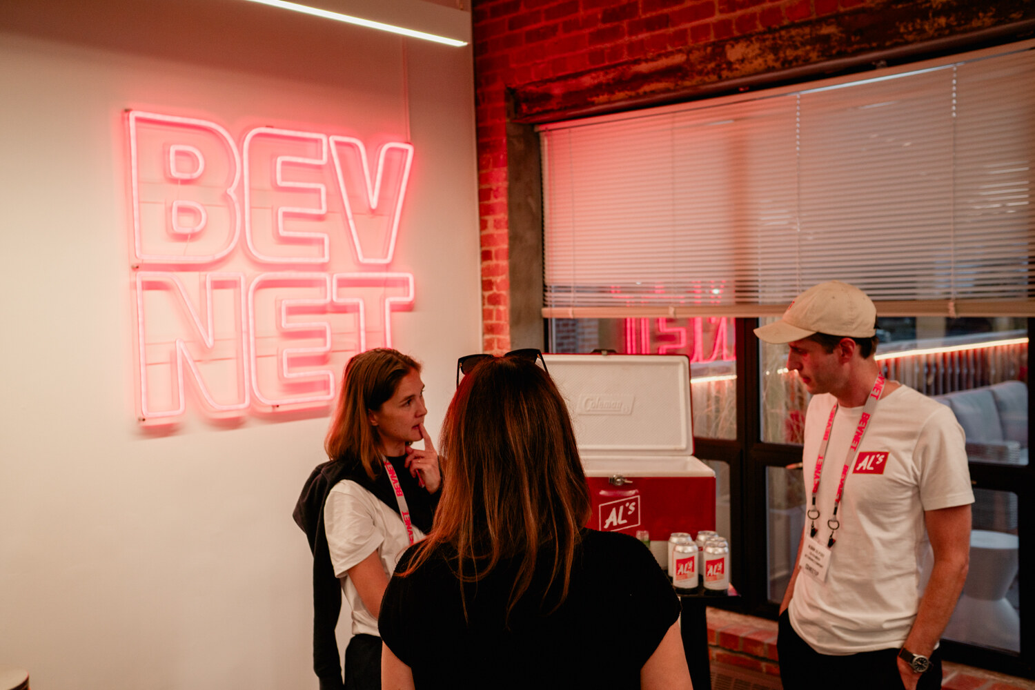 A red Al's cooler with white cans stands in front of a window beside a pink BEVNET neon sign while guests talk.