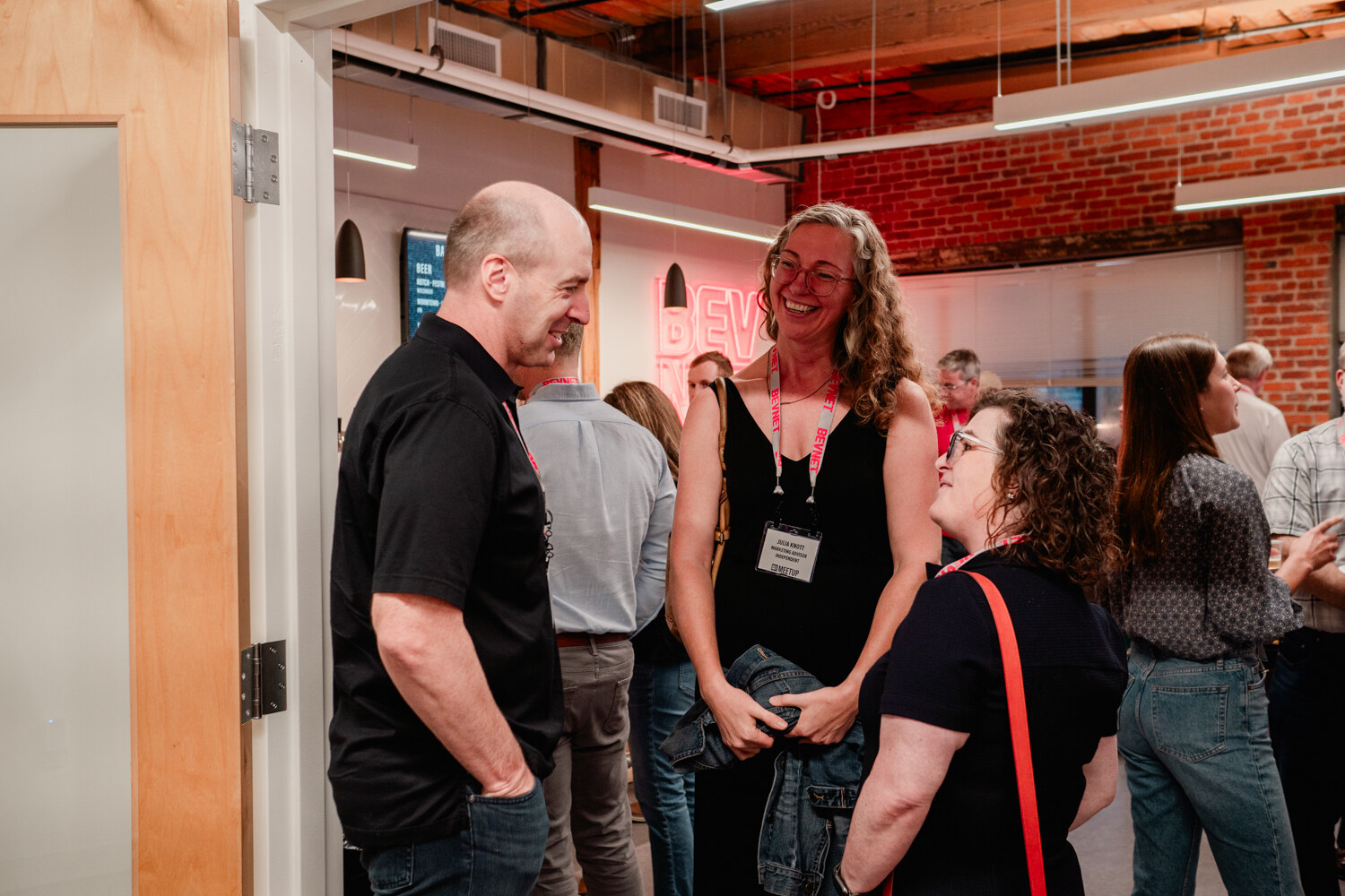 A man in a black polo and a woman in a black dress smile while talking with another woman beside the bar.