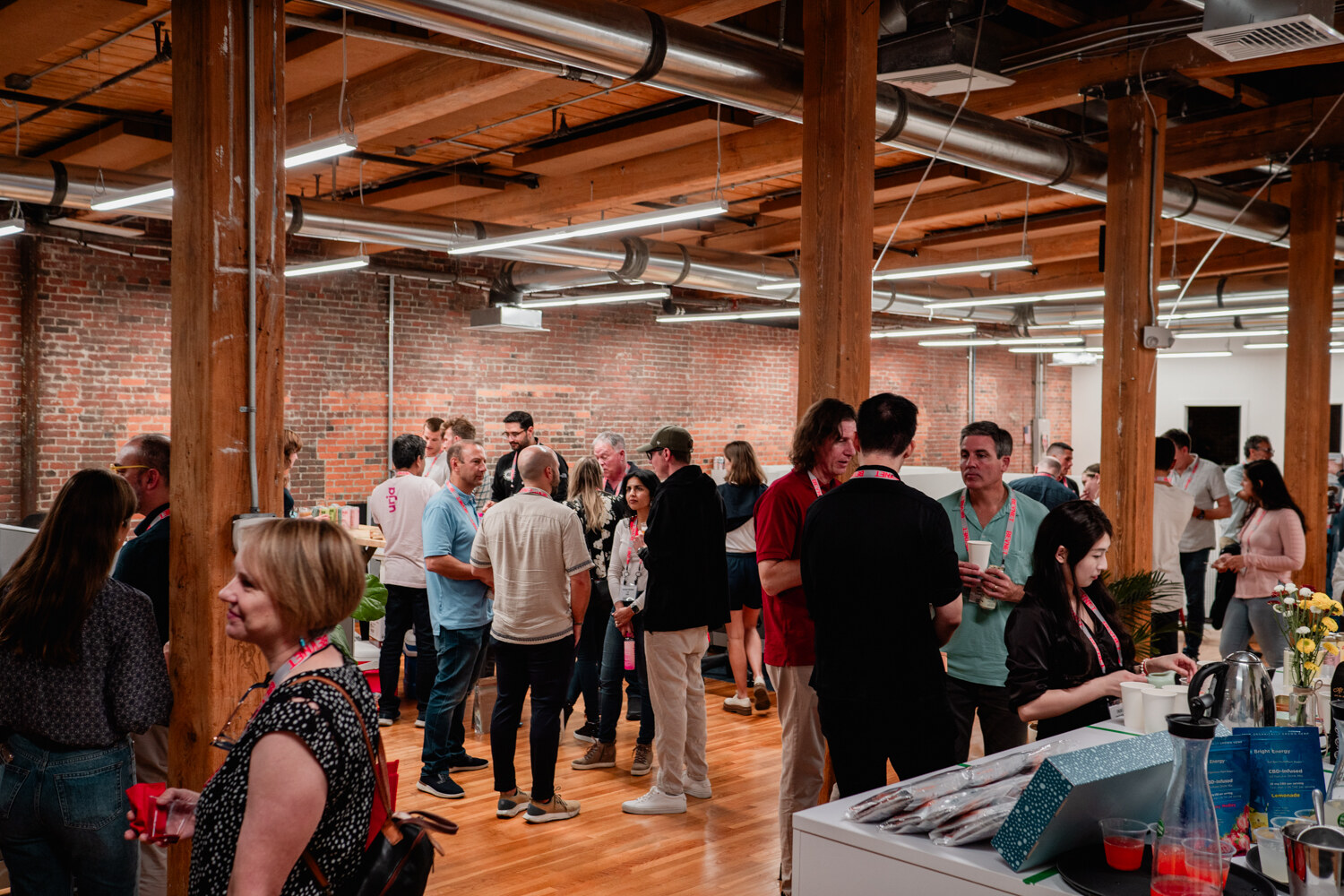 Wide view of dozens of guests talking across a brick-walled open office with wooden columns and tables.