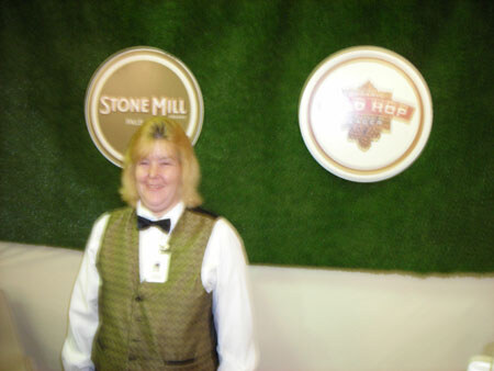 A blurry photo of a smiling woman with blonde hair in a green patterned vest, white shirt and black bow tie, standing in front of a green wall with a Stone Mill sign.