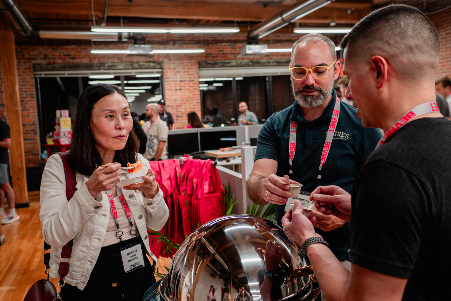 A woman in a white jacket samples a bite while two men in lanyards talk beside a chafing dish.