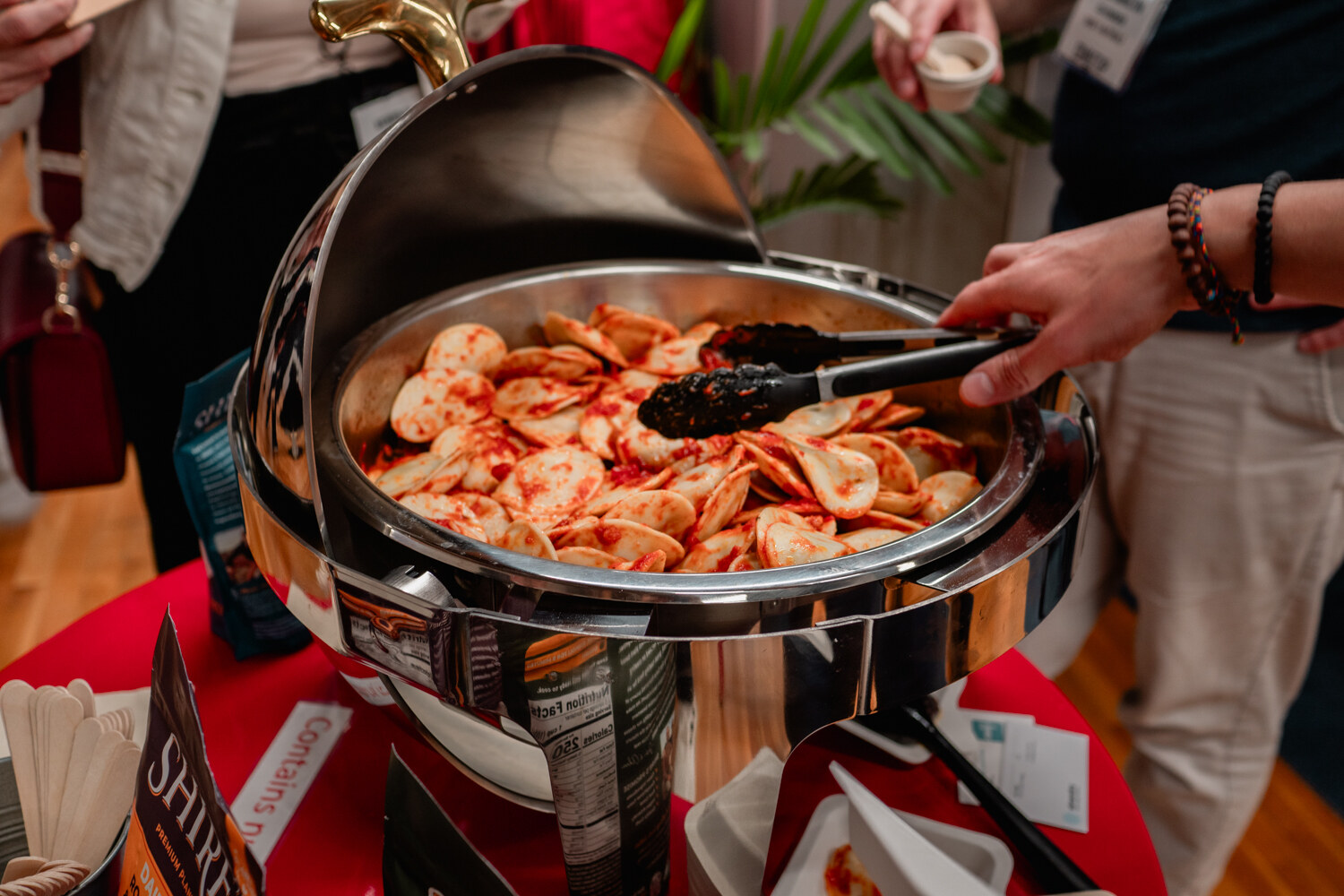 Close-up of a pair of tongs lifting pierogi in tomato sauce from a silver chafing dish.