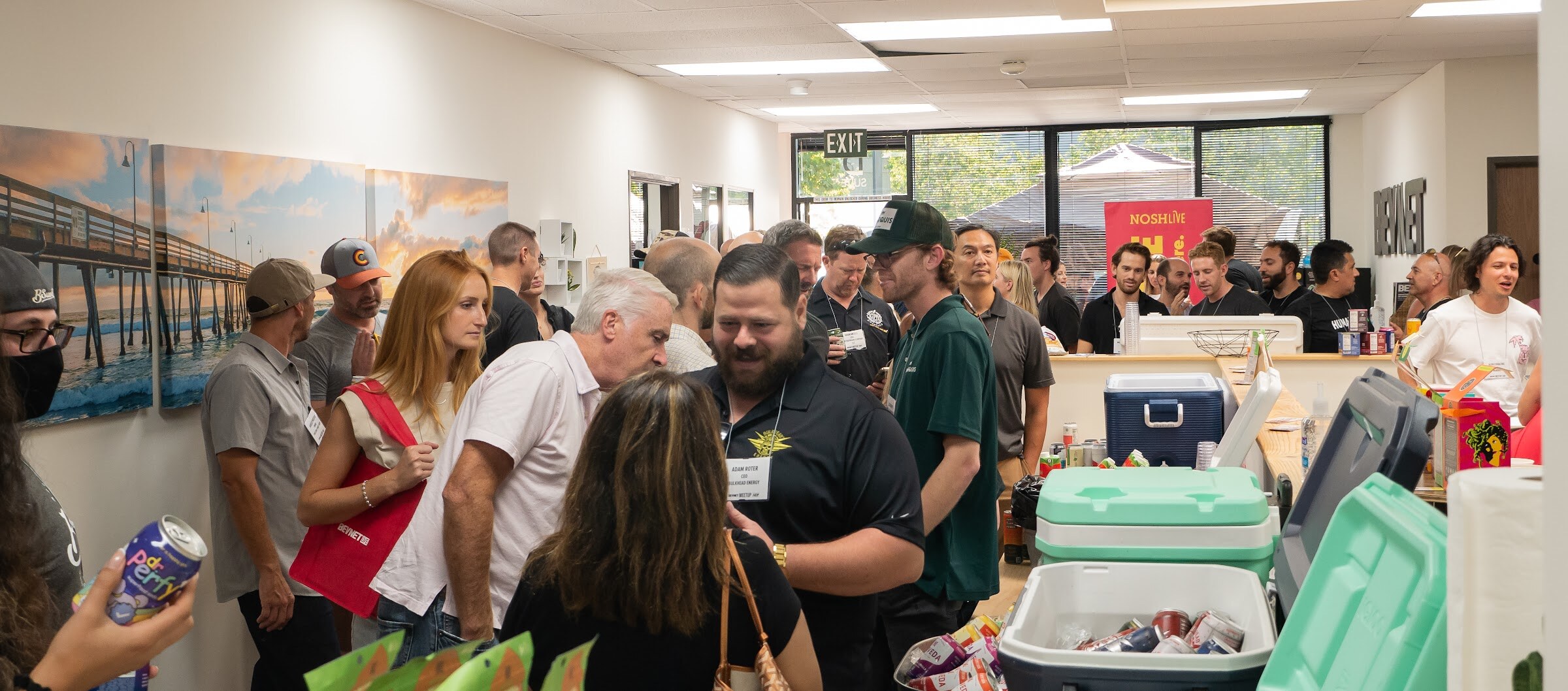 A crowded office with guests with name badges and drinks gathered around a counter with mint-green and white coolers, beneath wall photos of a pier at sunset and a red Nosh Live banner.