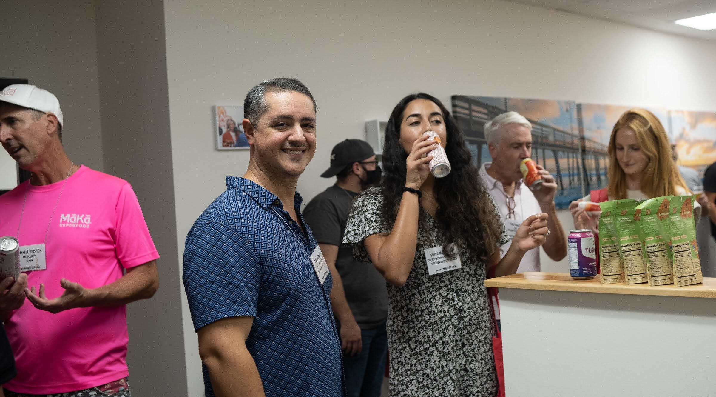 A man in a pink t-shirt, a smiling man in a blue patterned shirt, a woman in a floral dress drinking from a can, a man with gray hair and a woman with red hair stand talking beside a counter holding green snack packages.