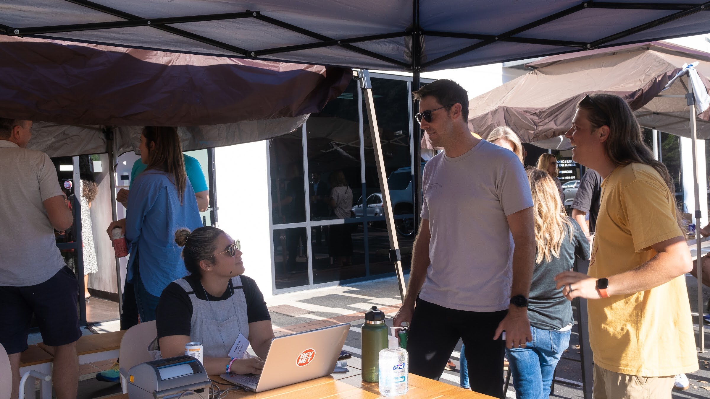 A woman in an apron sits at a table with a laptop and a label printer under a dark canopy while a man in a gray t-shirt and sunglasses and a man in a yellow t-shirt stand talking in front of her, with other guests behind.