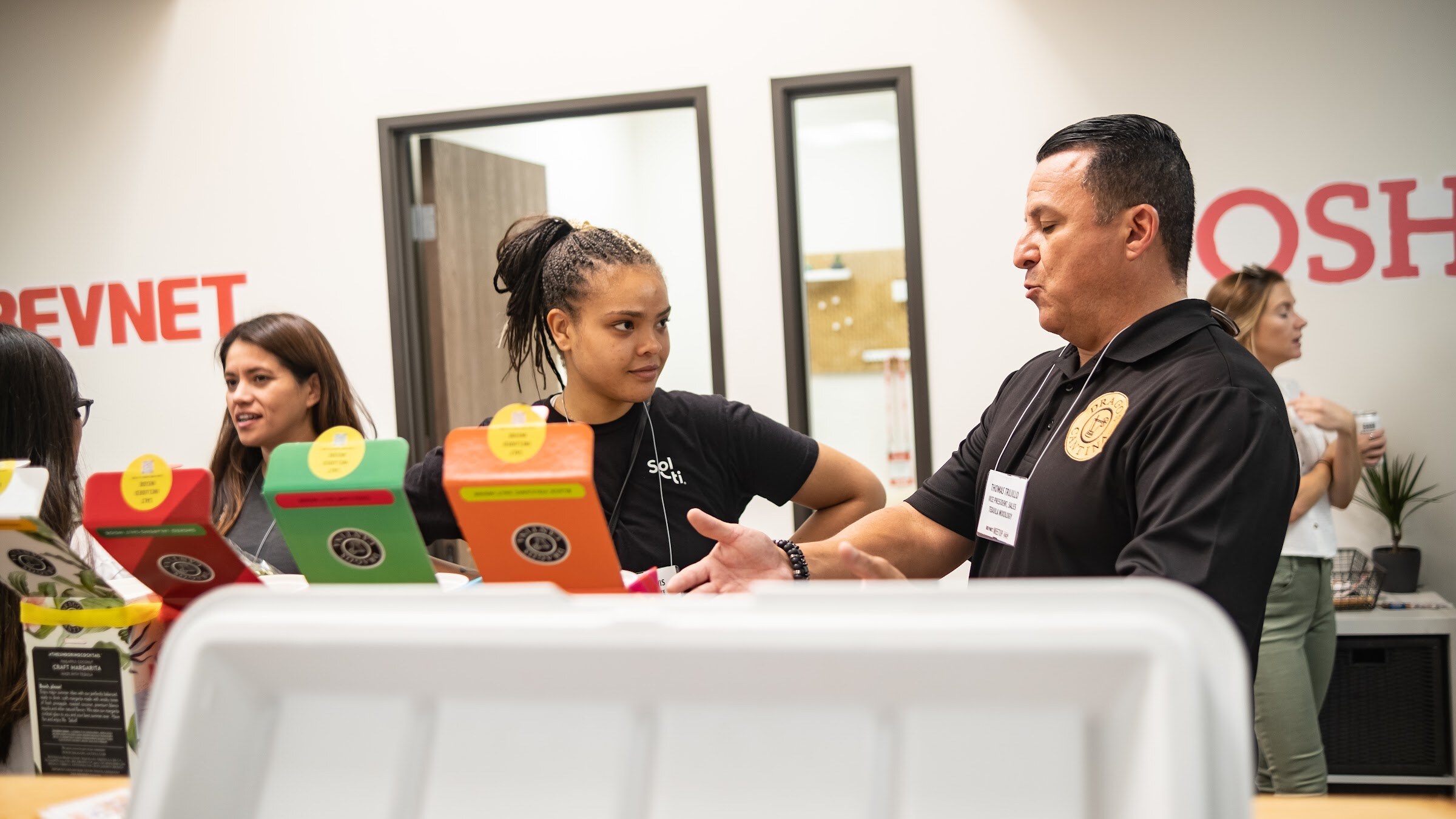 A man with a badge in a black polo gestures toward a woman with a ponytail and a woman with long hair behind a counter holding colorful boxes, in front of a white wall with red BEVNET and NOSH lettering.