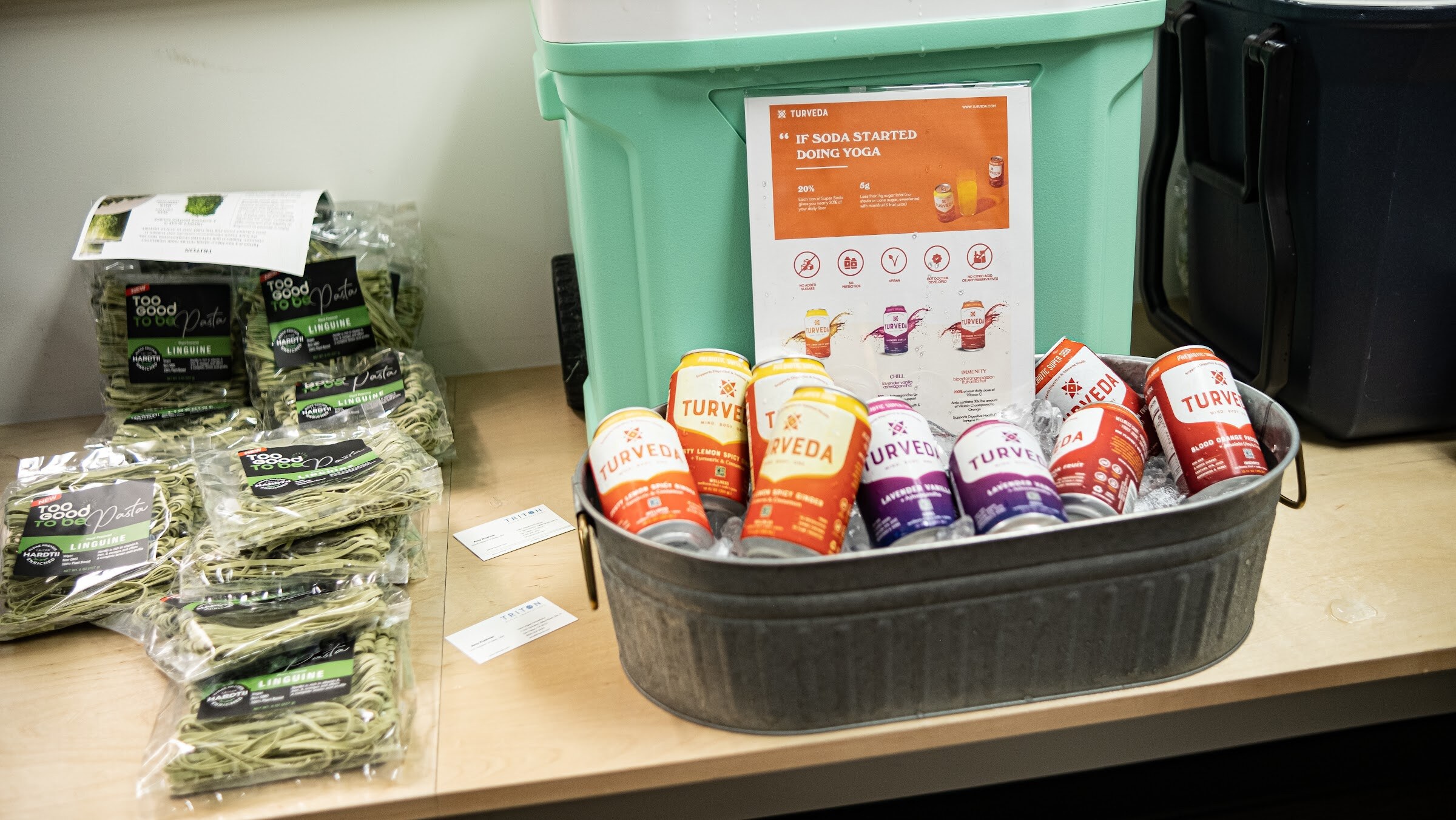 A galvanized tub of iced Turveda cans in orange, purple and red stands next to packages of linguine and a printed sign, beside a mint-green cooler on a wooden counter.