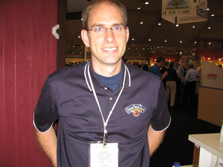 A smiling man with glasses wears a dark polo with a Stonyfield Farm logo and a show badge on a lanyard, standing in the expo hall.
