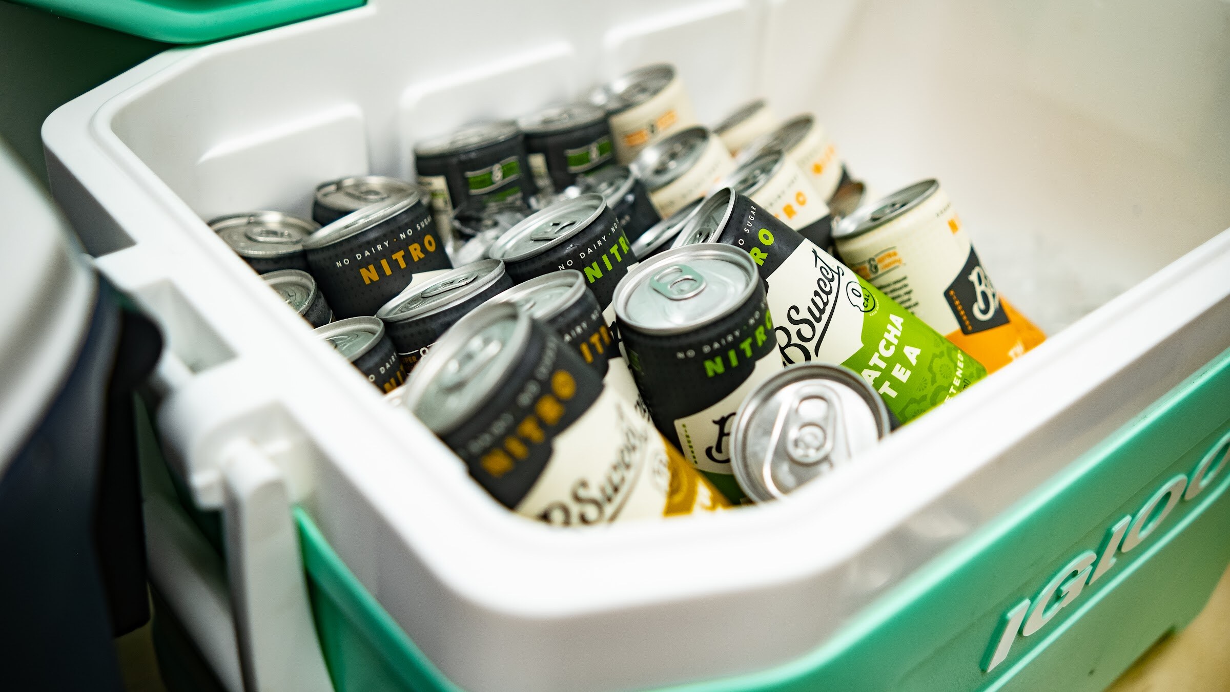 Black and white nitro cans including B Sweet Matcha Tea lie in a white and mint-green Igloo cooler.