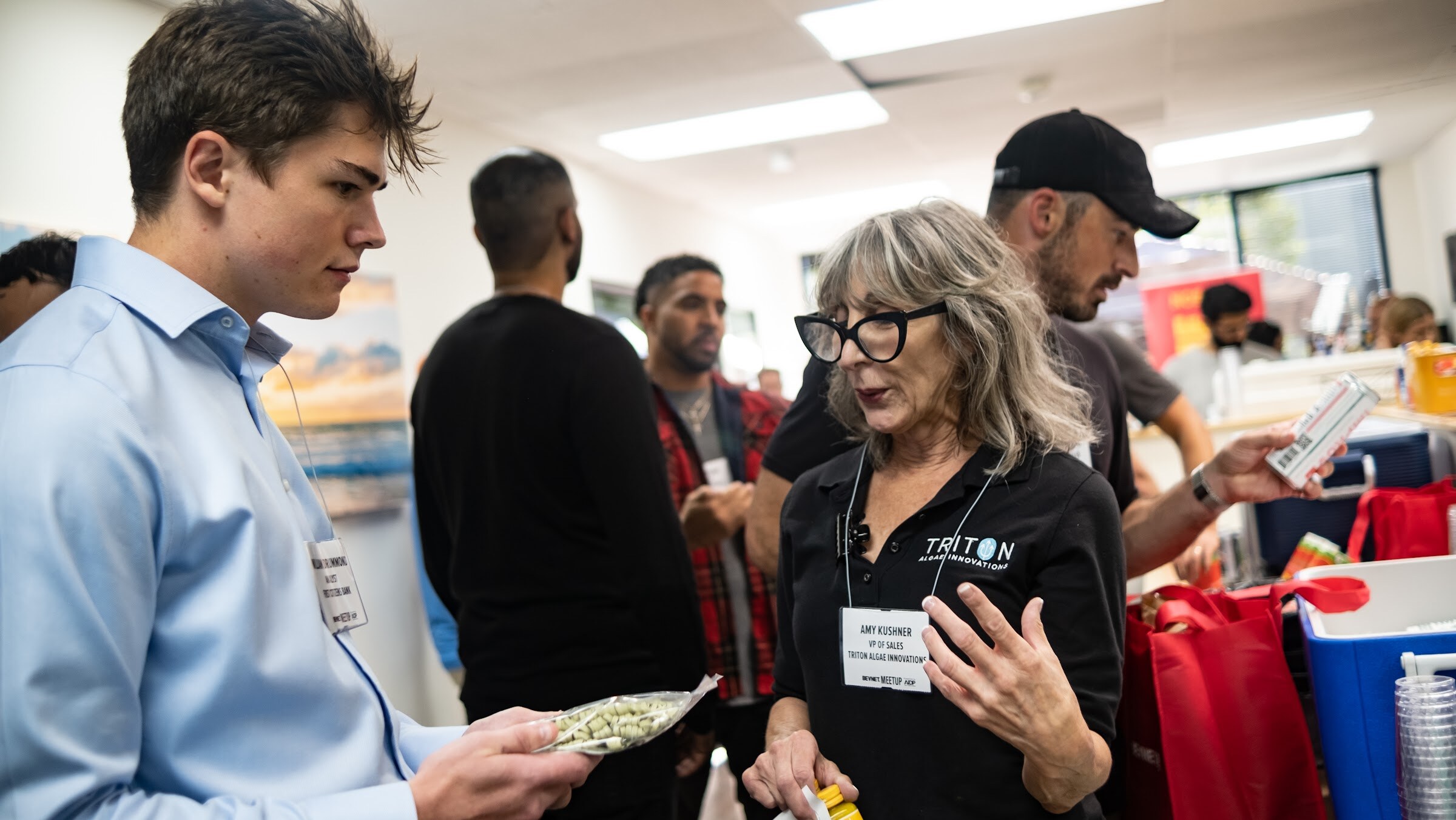 A woman with gray hair and black glasses in a black Triton polo talks with a young man in a light blue shirt who holds a package of green seeds, with guests, red tote bags and a cooler behind them.