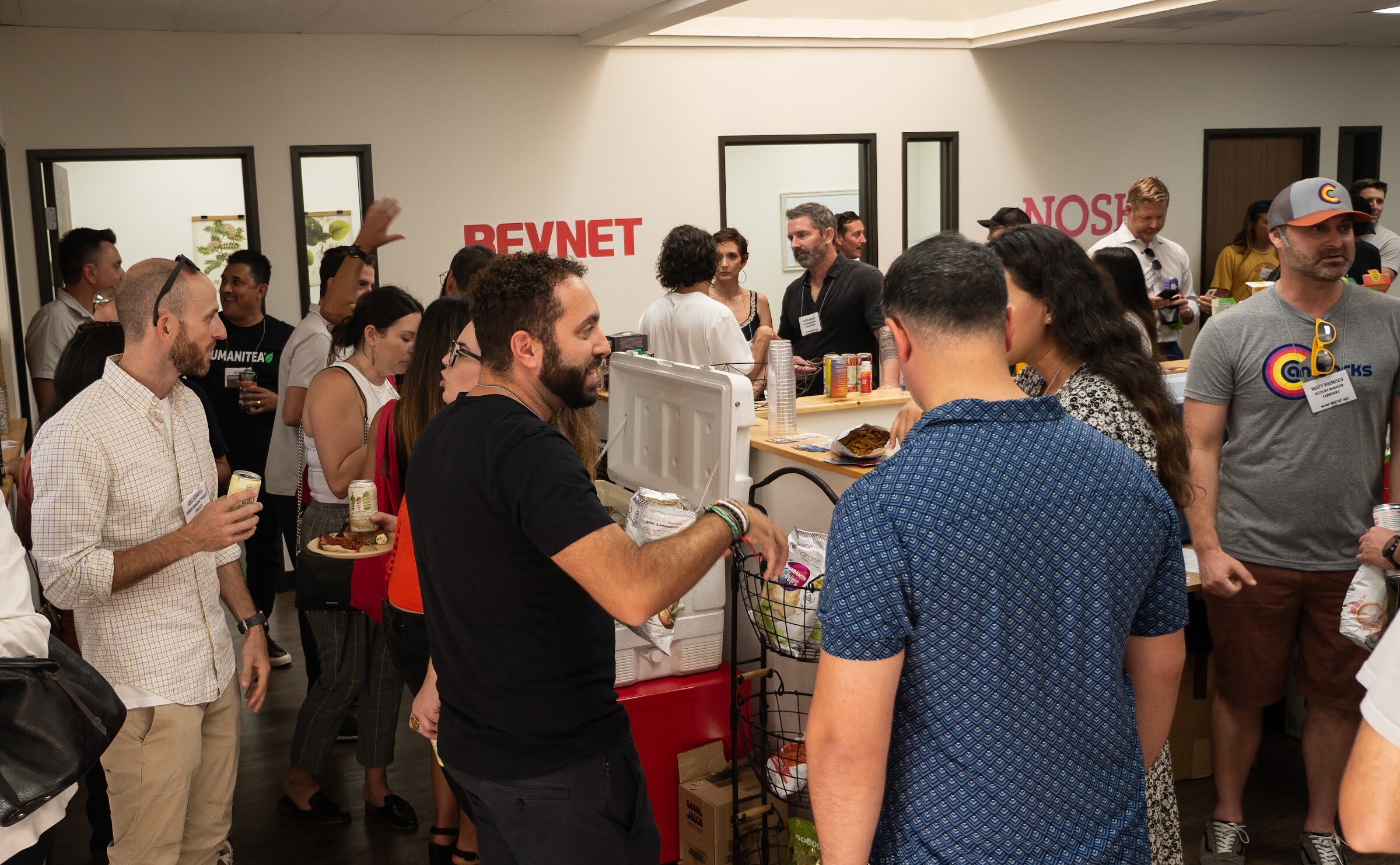 Guests with badges and plates talk around a counter with a cooler, including a bearded man in a black t-shirt and a man in a blue patterned shirt seen from behind, beneath red BEVNET and NOSH lettering on the wall.