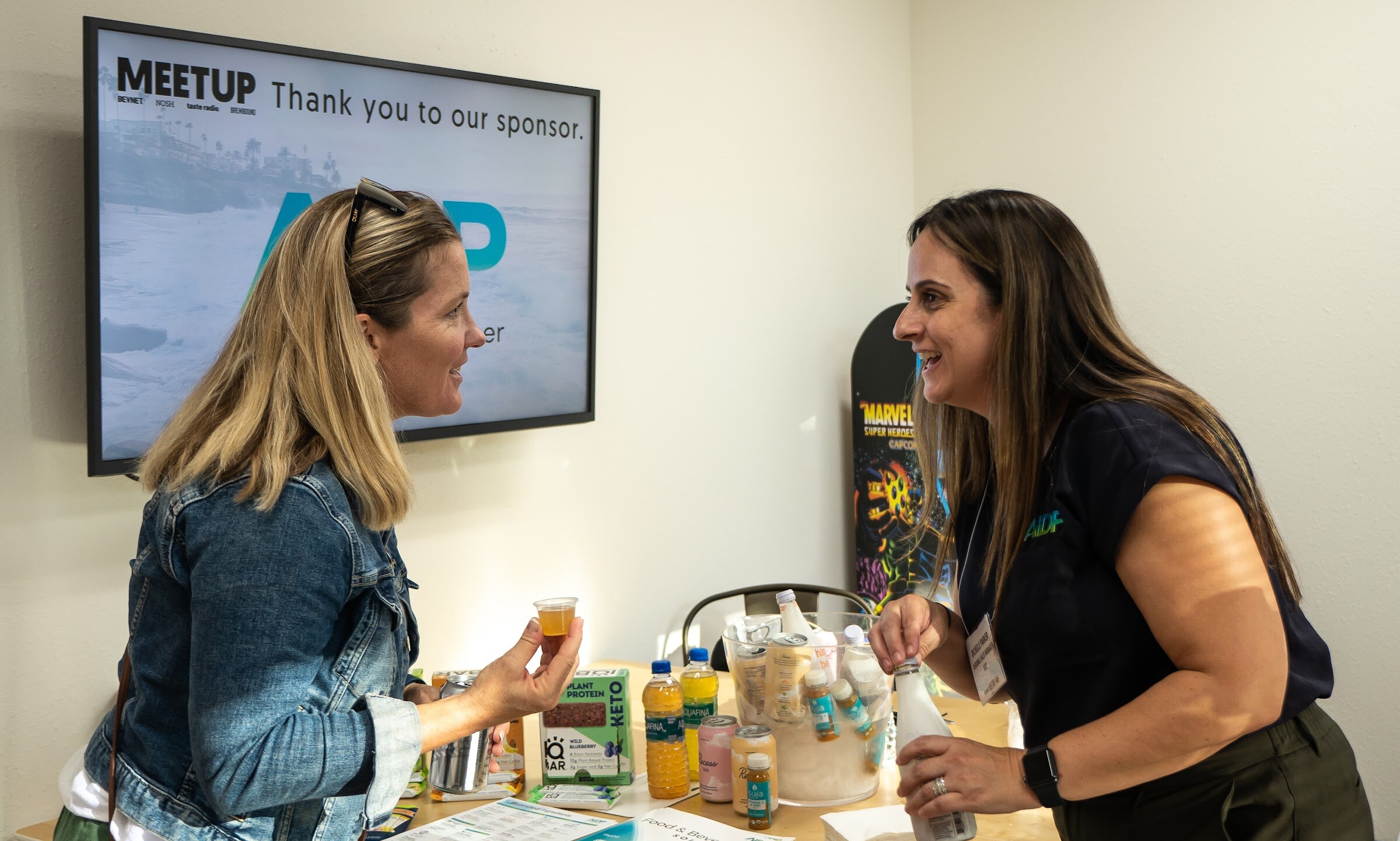 A woman with blonde hair in a denim jacket holds a small cup and talks with a smiling woman in a navy shirt, beside a table of bottles and cans and a wall screen reading Meetup, Thank you to our sponsor.