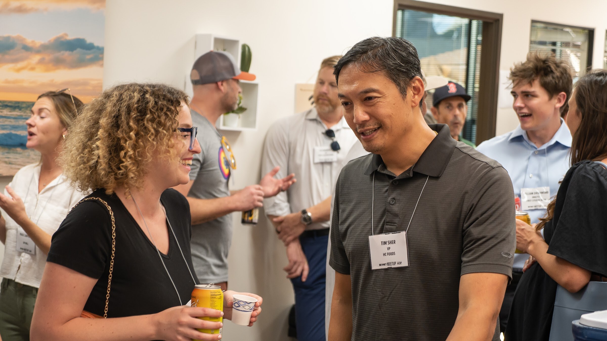 A woman with curly hair and glasses holding a yellow can and a cup talks with a smiling man in a gray polo with a name badge, with other guests and a beach sunset photo behind them.